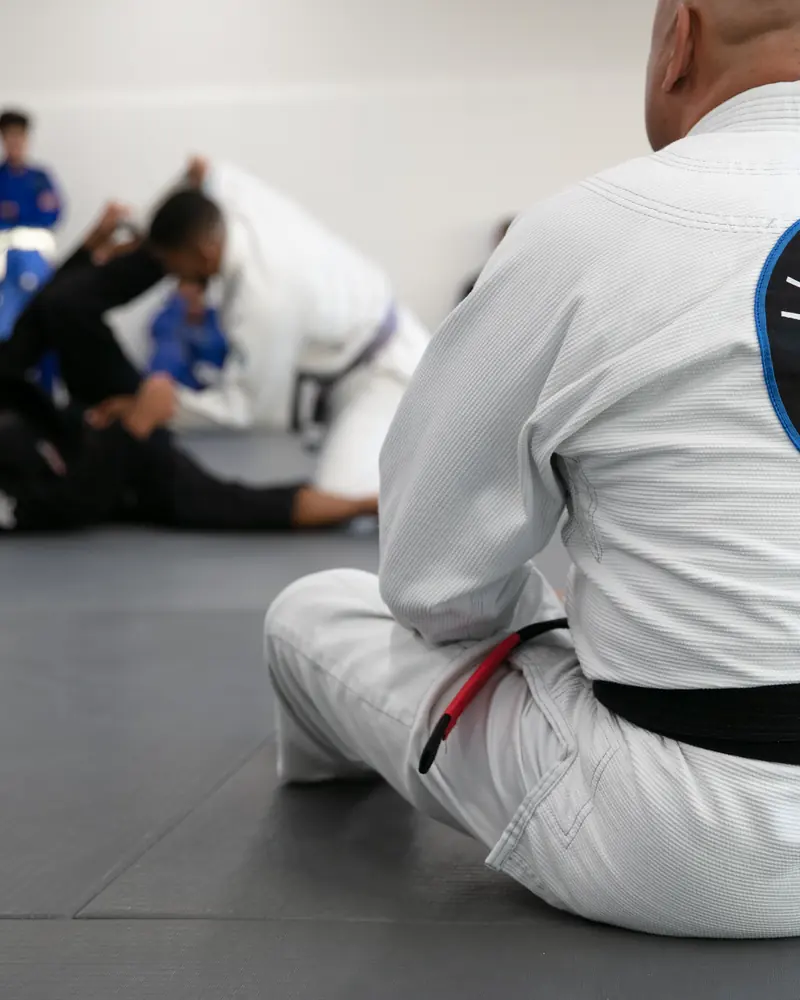 A student in a white gi sitting at the edge of the mat, watching the class