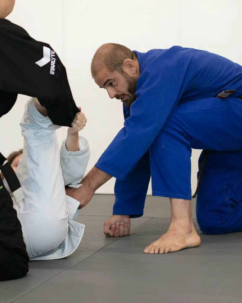 A coach in a blue gi kneeling beside two students, guiding a drill
