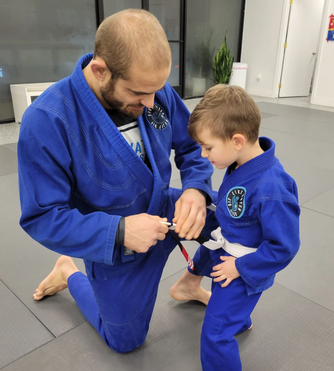 Mauricio kneeling to tie the white belt of a young student in a blue gi